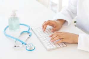 Closeup of female hands typing on computer keyboard, gel hand sanitizer and stethoscope on office desk. Online medical consultation and virus protection concept.