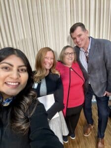 A group of 4 people posing for a selfie photo; from left to right visual descriptions: Indian American female with dark hair and black sweater, Caucasian female with strawberry blonde hair and black jacket over grey dress, Caucasian-presenting female blonde hair and dark rimmed glasses and red sweater set, Caucasian male with short blonde hair and blue suit jacket Caption: From left to right are Shree Thaker, Ashlie White, Patricia Nece, and Joe Nadglowski posing for a selfie photo at the end of the event. 