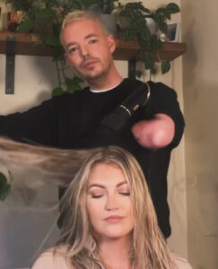 White male hairdresser uses a blow dryer on a blonde woman sitting in a chair.