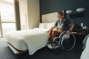 White female in a wheelchair shown in a hotel room next to the hotel bed.