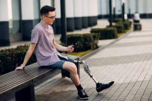 Young man sits on a bench outside and looks at his cellphone. He has one prosthetic leg.