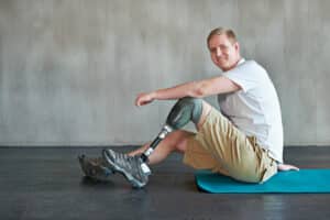 White male with blonde hair sits on a blue yoga mat with his elbow resting on his prosthetic leg. He is wearing a white tshirt and khaki shorts.