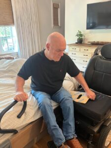 White male wheelchair user uses a transfer board to get onto a hotel bed.