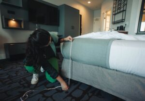 Female with long dark hair shown measuring bed height with a tape measure in a hotel room.