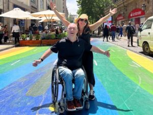 Man in a wheelchair and a woman behind him pose with their arms in the air. They are pictured on a multi-colored painted street.