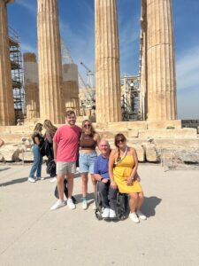 A man in a wheelchair with a woman on his lap and a young couple standing next to them. They are pictured in front of roman columns.