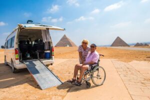Man in a wheelchair poses with a woman standing next to him. There is a wheelchair accessible rental van, equipped with a ramp, and its in front of the pyramids. 