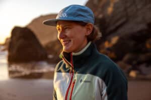 Nicole Ver Kuilen smiles while standing on a beach. She is wearing a fleece green and beige jacket and a blue cap.