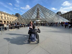 Wheelchair-user in front of the Louvre Museum. Lines of people waiting to get in are shown.