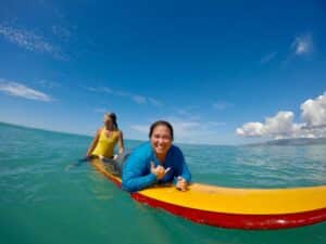 Woman lays on a longboard in the ocean and she is smiling. Another woman is positioned at the back of the board guiding her.